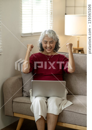 Asian senior woman celebrating online business success with joyful fist pump gesture at home achieving victory remote work lifestyle Asian senior woman celebrating online business success with joyful fist pump gesture at home achieving victory remote work lifestyle 139365806