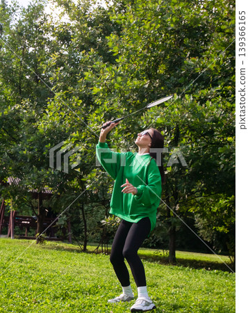 Young woman playing badminton in green park. Athletic adult holding racket during outdoor sports activity. Summer leisure, active lifestyle and healthy recreation in nature concept. Young woman playing badminton in green park. Athletic adult holding racket during outdoor sports activity. Summer leisure, active lifestyle and healthy recreation in nature concept. 139366165
