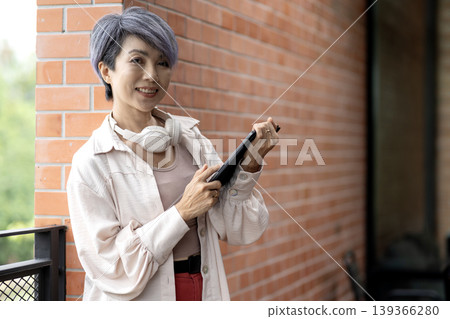 Asian adult woman smiling holding digital tablet browsing online content wearing headphones enjoying leisure time against brick wall urban outdoor setting Asian adult woman smiling holding digital tablet browsing online content wearing headphones enjoying leisure time against brick wall urban outdoor setting 139366280