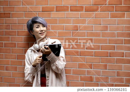 Asian adult woman using digital tablet and headphones against brick wall background enjoying modern technology lifestyle connection Asian adult woman using digital tablet and headphones against brick wall background enjoying modern technology lifestyle connection 139366281