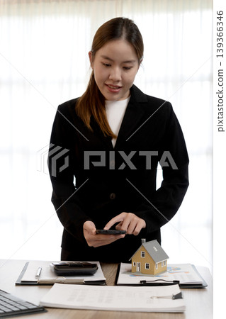 Asian adult professional woman real estate agent managing property business using smartphone desk property model investment home finance 139366346