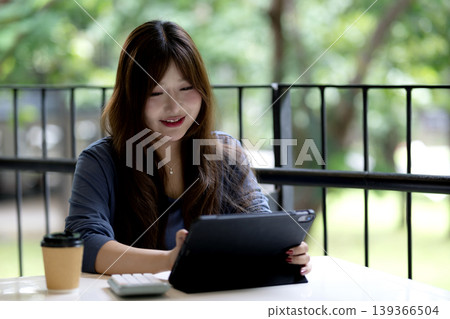 Young Asian woman performs digital tasks utilizing technology in a relaxing outdoor cafe environment fostering productivity and learning Young Asian woman performs digital tasks utilizing technology in a relaxing outdoor cafe environment fostering productivity and learning 139366504