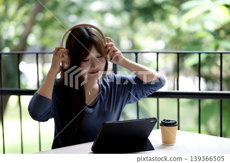 Asian adult woman uses wireless headphones and tablet device for remote work study or relaxed entertainment on an outdoor balcony surrounded by green nature Asian adult woman uses wireless headphones and tablet device for remote work study or relaxed entertainment on an outdoor balcony surrounded by green nature 139366505