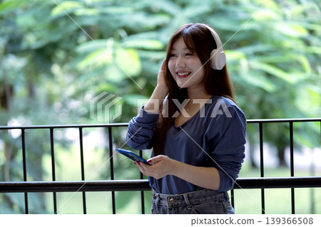 Asian woman architect enjoying music with headphones holding smartphone urban balcony green foliage leisure break design inspiration happy lifestyle relaxation moment 139366508