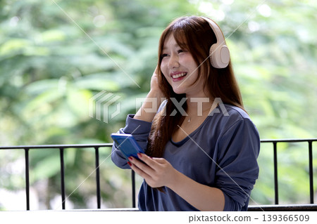 Asian woman adult enjoying music audio streaming outdoors with wireless headphones holding smartphone browsing content smiling relaxing on balcony nature background Asian woman adult enjoying music audio streaming outdoors with wireless headphones holding smartphone browsing content smiling relaxing on balcony nature background 139366509