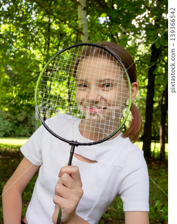 Young girl peeking through badminton racket in sunny park. Smiling child holding sports gear with green trees and outdoor nature background Young girl peeking through badminton racket in sunny park. Smiling child holding sports gear with green trees and outdoor nature background 139366542