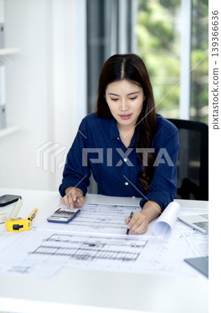Focused Asian architect reviews building designs making precise calculations on intricate plans at contemporary office workspace 139366636