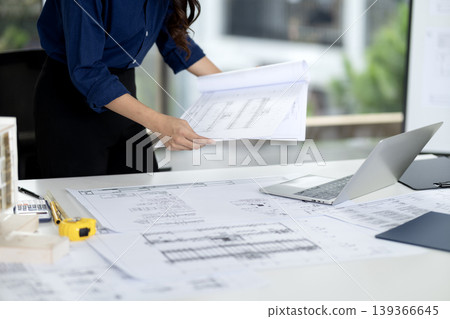 Adult woman architect reviews detailed building blueprints at bright modern office desk actively designing future construction projects Adult woman architect reviews detailed building blueprints at bright modern office desk actively designing future construction projects 139366645