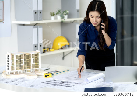 Adult Asian architect woman discussing project details on phone analyzing blueprints and building model in modern office workplace design engineering consultation Adult Asian architect woman discussing project details on phone analyzing blueprints and building model in modern office workplace design engineering consultation 139366712