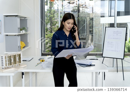 Asian adult architect discussing building plans on phone modern office design workspace professional woman collaborating project development urban construction 139366714