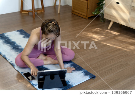 Asian adult woman practices yoga watching online tutorial tablet home workout promoting fitness wellness activity healthy lifestyle indoor space Asian adult woman practices yoga watching online tutorial tablet home workout promoting fitness wellness activity healthy lifestyle indoor space 139366967