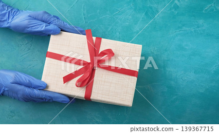 Quarantine surprise. Pandemic birthday. Hands in gloves holding wrapped gift box with red ribbon bow on blue textured background with free space. Quarantine surprise. Pandemic birthday. Hands in gloves holding wrapped gift box with red ribbon bow on blue textured background with free space. 139367715