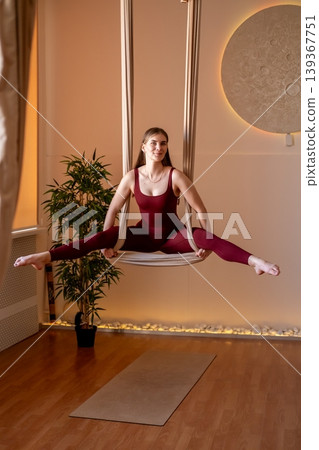 Yoga training. Split pose. Smiling woman in red sportswear hanging in sport swing hammock doing exercise for muscle stability in studio. Yoga training. Split pose. Smiling woman in red sportswear hanging in sport swing hammock doing exercise for muscle stability in studio. 139367751