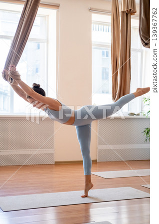 Flexibility training. Body balance. Woman in sportswear doing aerial yoga exercise standing on tiptoe leaning forward holding hammock in studio. Flexibility training. Body balance. Woman in sportswear doing aerial yoga exercise standing on tiptoe leaning forward holding hammock in studio. 139367762