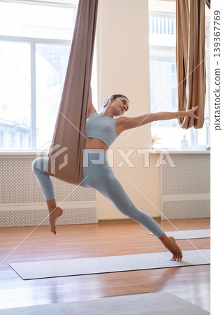 Fly yoga. Woman stretching. Young athlete doing gymnastics exercise in leaning pose supported by fabric hammock wrapped around leg in gym. Fly yoga. Woman stretching. Young athlete doing gymnastics exercise in leaning pose supported by fabric hammock wrapped around leg in gym. 139367769