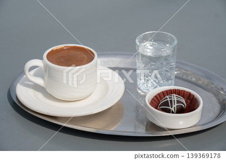 Turkish coffee in white cup on metal tray with glass of water and chocolate dessert, neutral background, minimal composition, soft light. Cafe service, beverage, food styling, hospitality, menu design Turkish coffee in white cup on metal tray with glass of water and chocolate dessert, neutral background, minimal composition, soft light. Cafe service, beverage, food styling, hospitality, menu design 139369178