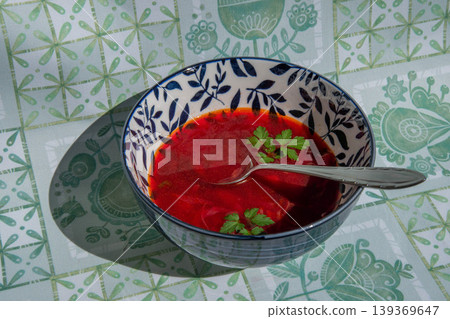 Ukrainian beetroot soup borscht poured into floral bowl over green cloth. Authentic borsch looks bright under sunlight with green herbs and metal spoon. Ukrainian beetroot soup borscht poured into floral bowl over green cloth. Authentic borsch looks bright under sunlight with green herbs and metal spoon. 139369647