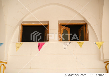 Mosque wall with arched opening and small wooden windows with shutters partially open and decorative hanging flags in foreground. Islamic architecture, facade detail, exterior design Mosque wall with arched opening and small wooden windows with shutters partially open and decorative hanging flags in foreground. Islamic architecture, facade detail, exterior design 139370646