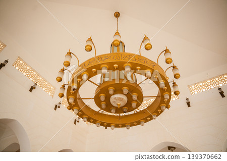 Large circular mosque chandelier with Arabic calligraphy inscriptions and multiple hanging lamps mounted under a white domed ceiling. Islamic religion, architecture, interior lighting Large circular mosque chandelier with Arabic calligraphy inscriptions and multiple hanging lamps mounted under a white domed ceiling. Islamic religion, architecture, interior lighting 139370662