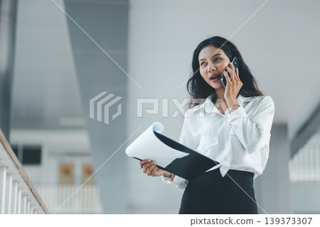 Professional woman speaking on a smartphone while reviewing documents on a clipboard in a bright office corridor, ideal for business communication, client support, multitasking, and planning. Professional woman speaking on a smartphone while reviewing documents on a clipboard in a bright office corridor, ideal for business communication, client support, multitasking, and planning. 139373307