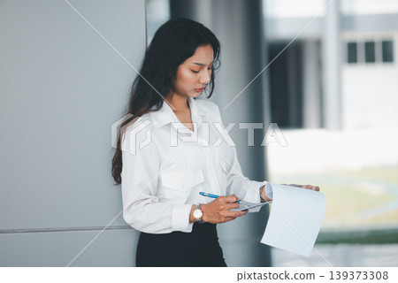 Professional woman checking paperwork with pen while standing in a modern office, suitable for corporate administration, report review, business planning, concentration, and workplace routine. 139373308