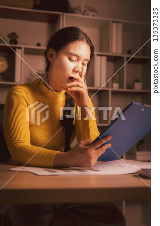 Sleepy young Asian woman yawning while reviewing clipboard documents in cozy home office at night, illustrating paperwork fatigue, overtime stress, deadline pressure and evening workload. Sleepy young Asian woman yawning while reviewing clipboard documents in cozy home office at night, illustrating paperwork fatigue, overtime stress, deadline pressure and evening workload. 139373385