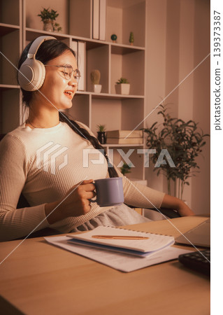 Smiling young Asian woman wearing headphones and holding coffee while relaxing by laptop in cozy home office at night, representing remote work break, online learning, comfort and digital lifestyle. Smiling young Asian woman wearing headphones and holding coffee while relaxing by laptop in cozy home office at night, representing remote work break, online learning, comfort and digital lifestyle. 139373387