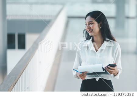 Smiling professional woman holding a smartphone, documents, and pen in an office walkway, showing successful communication, confidence, client coordination, and corporate productivity. Smiling professional woman holding a smartphone, documents, and pen in an office walkway, showing successful communication, confidence, client coordination, and corporate productivity. 139373388