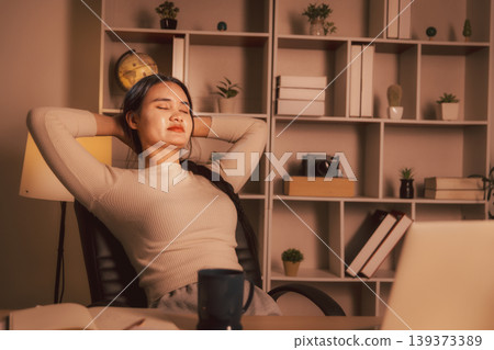 Woman relaxing with eyes closed and hands behind head in cozy home office at night, illustrating work break, stress relief, remote work burnout recovery, wellness and calm digital lifestyle. 139373389