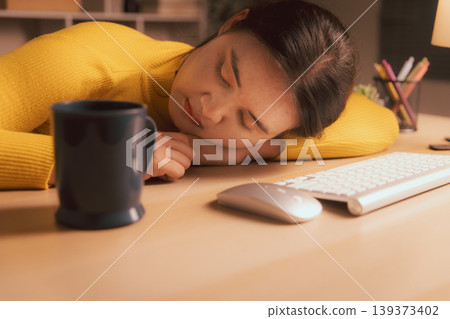 Exhausted young Asian woman sleeping at desk beside keyboard and coffee in cozy home office at night, illustrating burnout, overtime, work fatigue, deadline stress and late night productivity loss. Exhausted young Asian woman sleeping at desk beside keyboard and coffee in cozy home office at night, illustrating burnout, overtime, work fatigue, deadline stress and late night productivity loss. 139373402