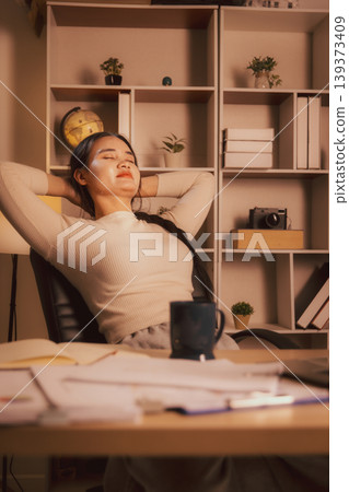Woman relaxing with eyes closed and hands behind head in cozy home office at night, illustrating work break, stress relief, remote work burnout recovery, wellness and calm digital lifestyle. Woman relaxing with eyes closed and hands behind head in cozy home office at night, illustrating work break, stress relief, remote work burnout recovery, wellness and calm digital lifestyle. 139373409