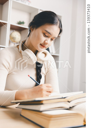 Young Asian woman studying at home office, writing notes with books, laptop and headphones on desk. Online learning, remote education, exam preparation, focus and productivity concept. Young Asian woman studying at home office, writing notes with books, laptop and headphones on desk. Online learning, remote education, exam preparation, focus and productivity concept. 139373410