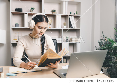 Young Asian woman studying at home with headphones, reading a book, taking notes and using a laptop at desk. Online learning, focused education, remote study and productivity concept. 139373450