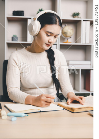 Young Asian woman studying at home with headphones, reading a book, taking notes and using a laptop at desk. Online learning, focused education, remote study and productivity concept. Young Asian woman studying at home with headphones, reading a book, taking notes and using a laptop at desk. Online learning, focused education, remote study and productivity concept. 139373451