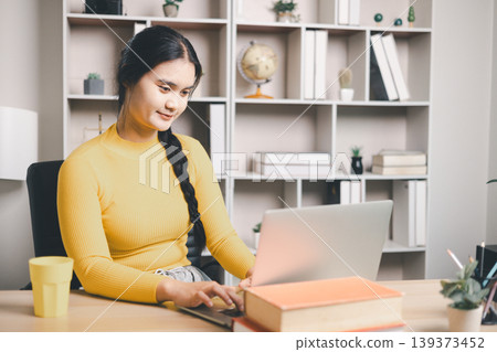 Young Asian woman using laptop at home office desk with books and coffee, studying online or working remotely. Smiling student in modern workspace, e-learning, productivity and lifestyle concept. Young Asian woman using laptop at home office desk with books and coffee, studying online or working remotely. Smiling student in modern workspace, e-learning, productivity and lifestyle concept. 139373452