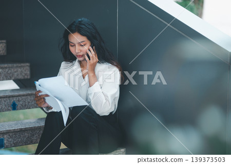 Businesswoman talking on a smartphone while reading reports on office stairs, showing mobile communication, multitasking, document review, problem solving, and professional productivity. Businesswoman talking on a smartphone while reading reports on office stairs, showing mobile communication, multitasking, document review, problem solving, and professional productivity. 139373503