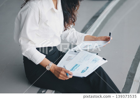Businesswoman reviewing printed charts and market reports while seated with a clipboard, showing financial analysis, strategic planning, document management, and focused office work. Businesswoman reviewing printed charts and market reports while seated with a clipboard, showing financial analysis, strategic planning, document management, and focused office work. 139373535