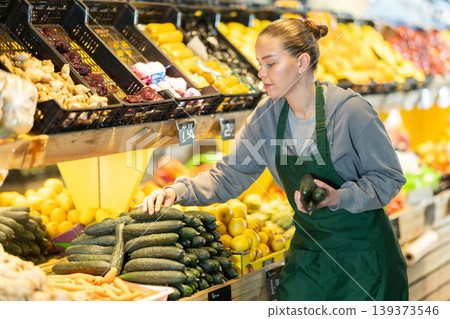 saleswoman stands near a display case and checks the quality of cucumbers saleswoman stands near a display case and checks the quality of cucumbers 139373546