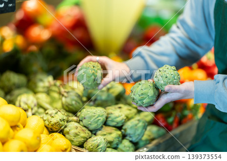 Close-up of salesperson hand put ripe artichokes on grocery supermarket display Close-up of salesperson hand put ripe artichokes on grocery supermarket display 139373554
