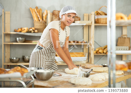 Young female baker kneading dough on flour-dusted table in artisan bakery Young female baker kneading dough on flour-dusted table in artisan bakery 139373880