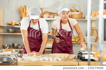 Two young bakers prepare dough in kitchen of bakery - together they knead dough, cut it with knife and weigh it on scales Two young bakers prepare dough in kitchen of bakery - together they knead dough, cut it with knife and weigh it on scales 139374090