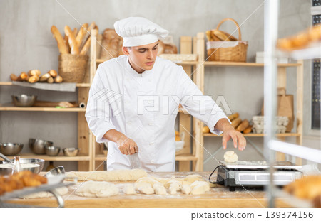 Young male baker cutting and weighing dough on scales in bakehouse Young male baker cutting and weighing dough on scales in bakehouse 139374156