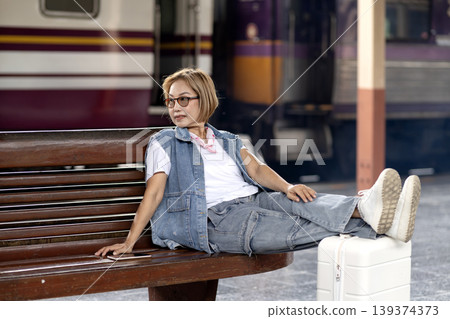 Asian woman traveler rests platform seat awaiting train departure journey travel adventure suitcase phone near hand vacation relaxation 139374373