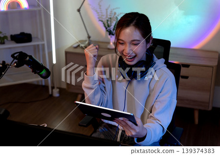 Asian adult woman gamer streaming celebrates victory playing online game on tablet broadcasting from modern home studio setup colorful neon lights Asian adult woman gamer streaming celebrates victory playing online game on tablet broadcasting from modern home studio setup colorful neon lights 139374383