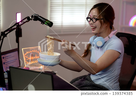 Asian woman gamer streamer eating instant noodles chopsticks laughing joyfully while live video stream broadcast online home room modern technology gaming lifestyle fun content creator 139374385