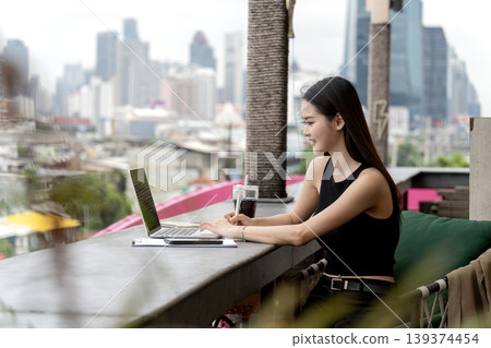 Asian young adult woman working on laptop at city rooftop cafe productive professional remote work lifestyle digital nomad urban setting Asian young adult woman working on laptop at city rooftop cafe productive professional remote work lifestyle digital nomad urban setting 139374454