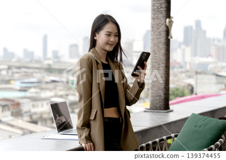 Asian adult woman using smartphone managing business outdoor balcony urban skyline working remotely digital nomad professional modern city view technology connectivity lifestyle Asian adult woman using smartphone managing business outdoor balcony urban skyline working remotely digital nomad professional modern city view technology connectivity lifestyle 139374455