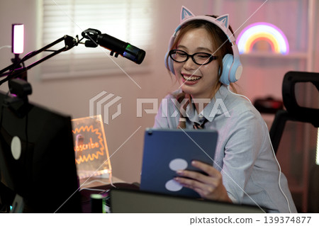 Asian adult woman streaming gaming podcast content creation using tablet microphone headphones with joy at home studio personal room Asian adult woman streaming gaming podcast content creation using tablet microphone headphones with joy at home studio personal room 139374877