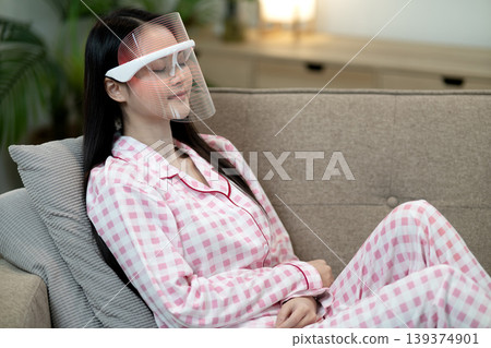 Asian adult woman applying LED light therapy mask home for skin care beauty treatment facial rejuvenation anti aging relaxation living room couch Asian adult woman applying LED light therapy mask home for skin care beauty treatment facial rejuvenation anti aging relaxation living room couch 139374901