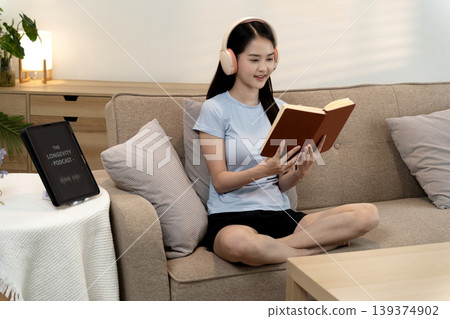 Asian young adult woman wearing headphones reading book relaxing on sofa in modern cozy living room enjoying quiet leisure time learning wellness and longevity Asian young adult woman wearing headphones reading book relaxing on sofa in modern cozy living room enjoying quiet leisure time learning wellness and longevity 139374902
